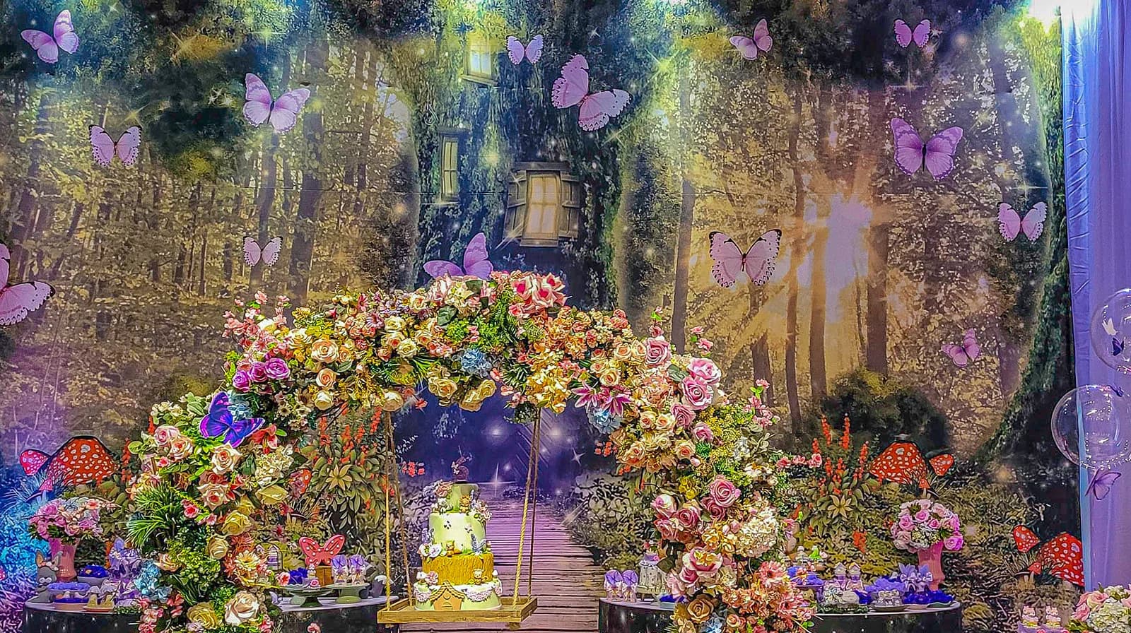 Decoração de floresta encantada com arco de flores, borboletas e painel de árvores.
