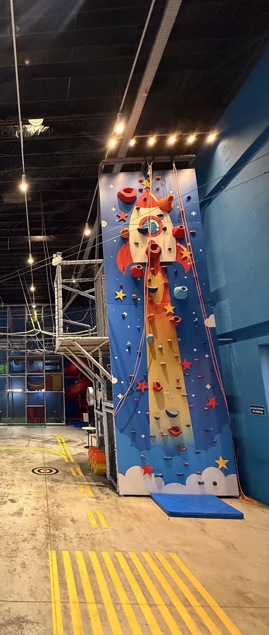 Parede de escalada azul com um foguete desenhado, ao lado do kid play.