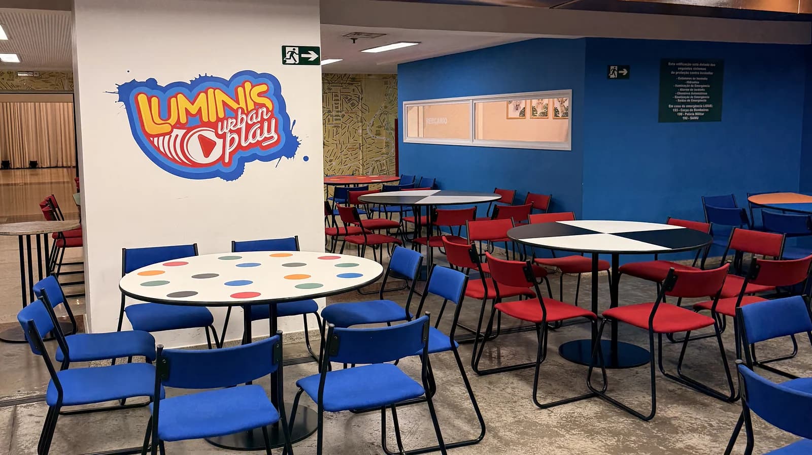 Salão de mesas redondas com cadeiras azuis e vermelhas e o logo do Luminis na parede.